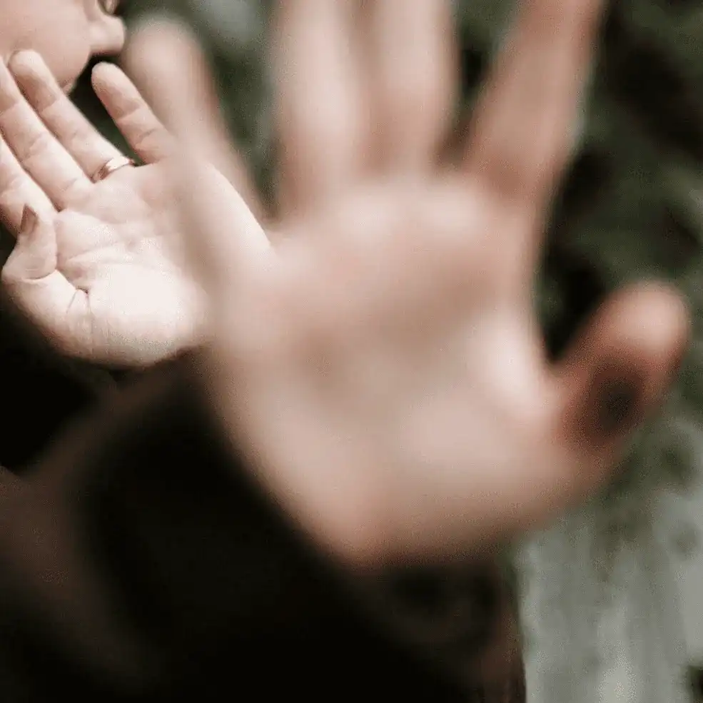 A person holding their hand up toward the camera, partially blocking the lens, with a blurred background.